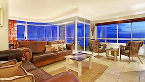 A modern living room with a leather sofa, a wooden coffee table, wicker chairs, and large windows offering a view of the sky. - Ocean View C602 bt HostAgents (Cape Town)