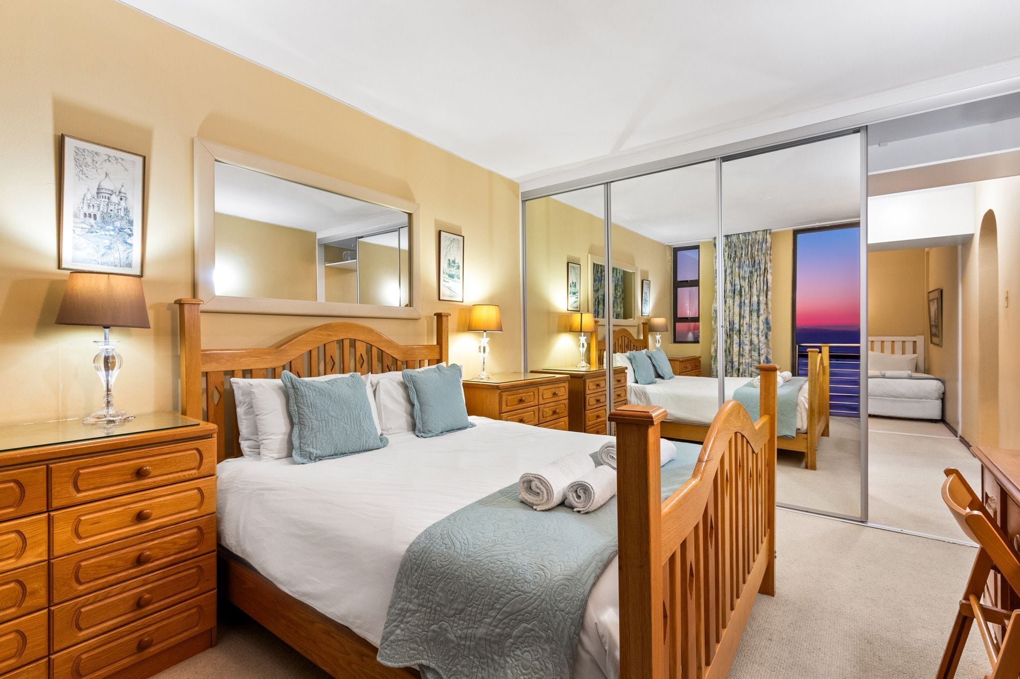 A hotel room with a large bed, wooden headboard, a dresser with drawers, a mirror, and a view of the outside through a glass door.