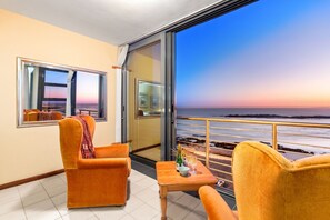 A balcony with two orange chairs, a small table with a bottle and glasses, and a view of the ocean at sunset. - Sand and See 302 (Cape Town)