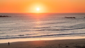 Ein Strandbild mit Sonnenuntergang über dem Meer, eine Person steht im Sand, und es liegen Felsen im Wasser.