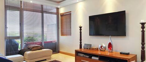 A modern living room with a sofa, a flat-screen TV mounted on the wall, a wooden entertainment unit, and large windows with blinds.