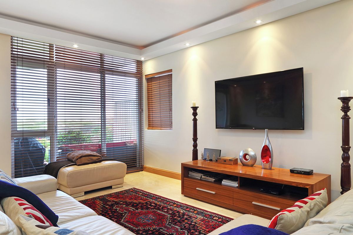 Un salon moderne comprenant un canapé, un téléviseur à écran plat fixé au mur, un meuble de rangement en bois et de grandes fenêtres équipées de stores.