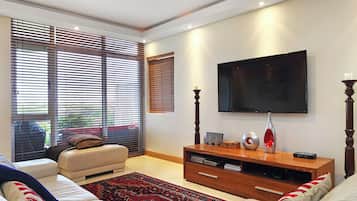 A modern living room with a sofa, a flat-screen TV mounted on the wall, a wooden entertainment unit, and large windows with blinds.