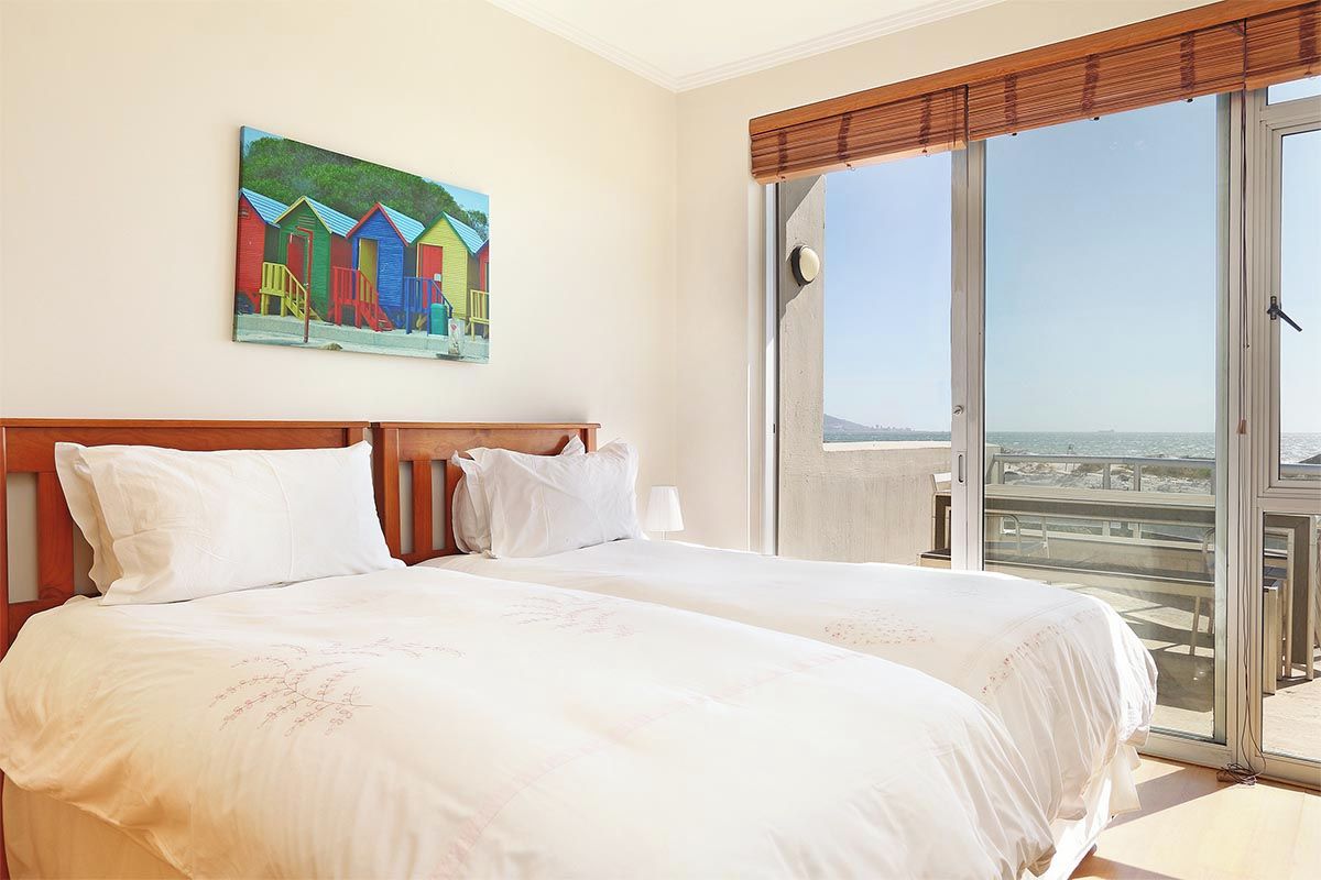 Une chambre d’hôtel avec deux lits, une grande fenêtre donnant sur la plage et un tableau représentant des cabanes de plage aux couleurs vives accroché au mur.
