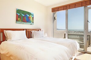A hotel room with two beds, a large window with a view of the beach, and a painting of colorful beach huts on the wall. - Dolphin Beach E5 (Cape Town)