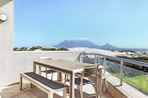 A balcony with a table and chairs, overlooking a beach and a mountain. - Dolphin Beach E5 (Cape Town)