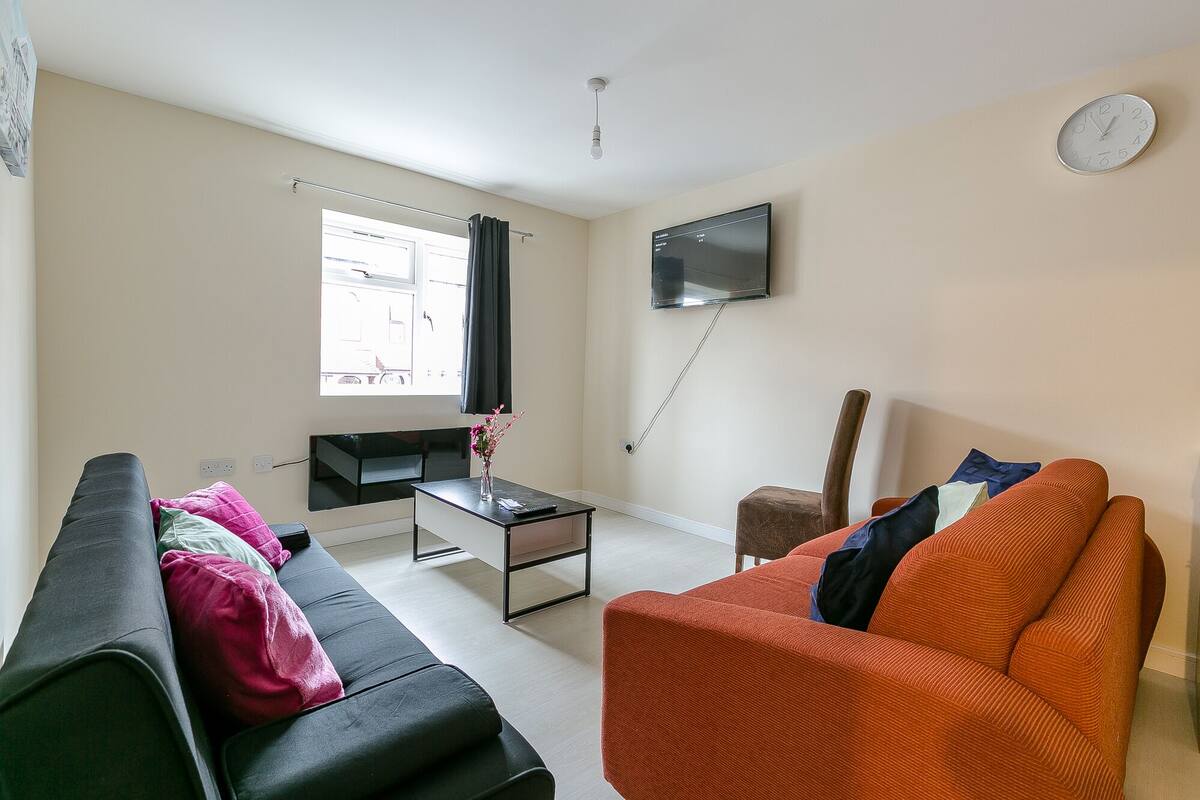 A living room with a sofa, a coffee table, a TV mounted on the wall, and a clock.