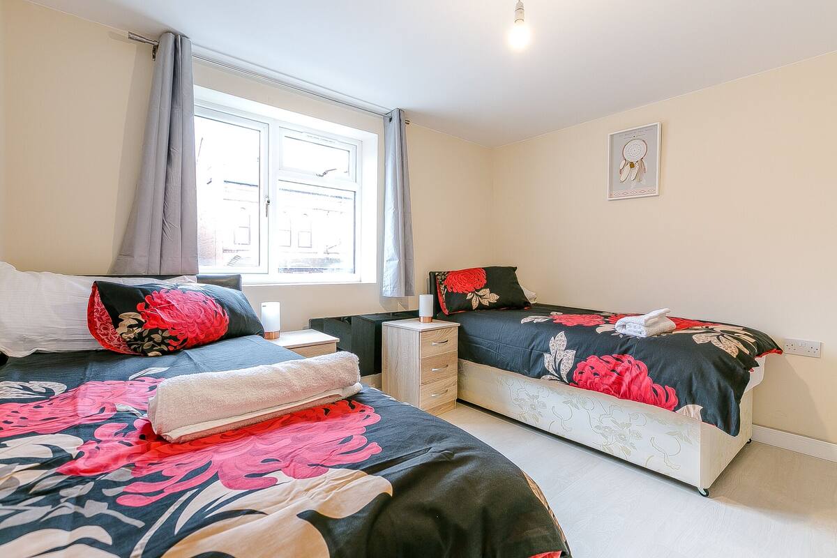A bedroom with two beds, a nightstand, a bedside table, a window with curtains, and a wall-mounted artwork.