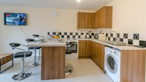 A compact kitchen with wooden cabinets, a built-in washing machine, a gas stove, and a bar stool. - Kynsparks Luxury Apartments (Birmingham)