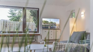 Un salon moderne comprenant un canapé, une table basse, un téléviseur et donnant sur un quartier résidentiel grâce à de grandes fenêtres.