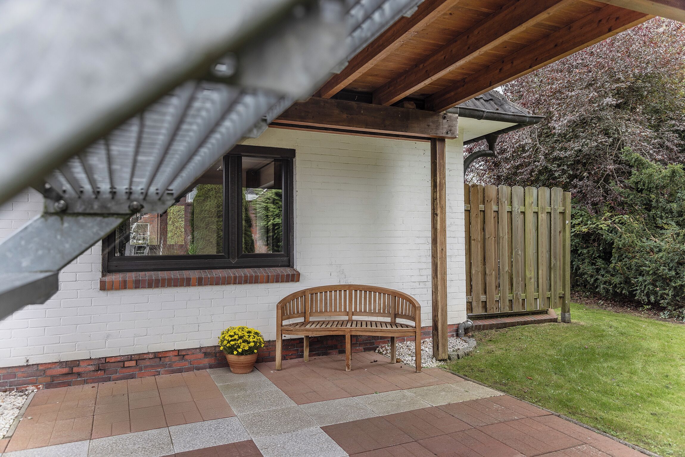 A wooden bench under a covered patio area with a white brick wall and a wooden fence in the background.