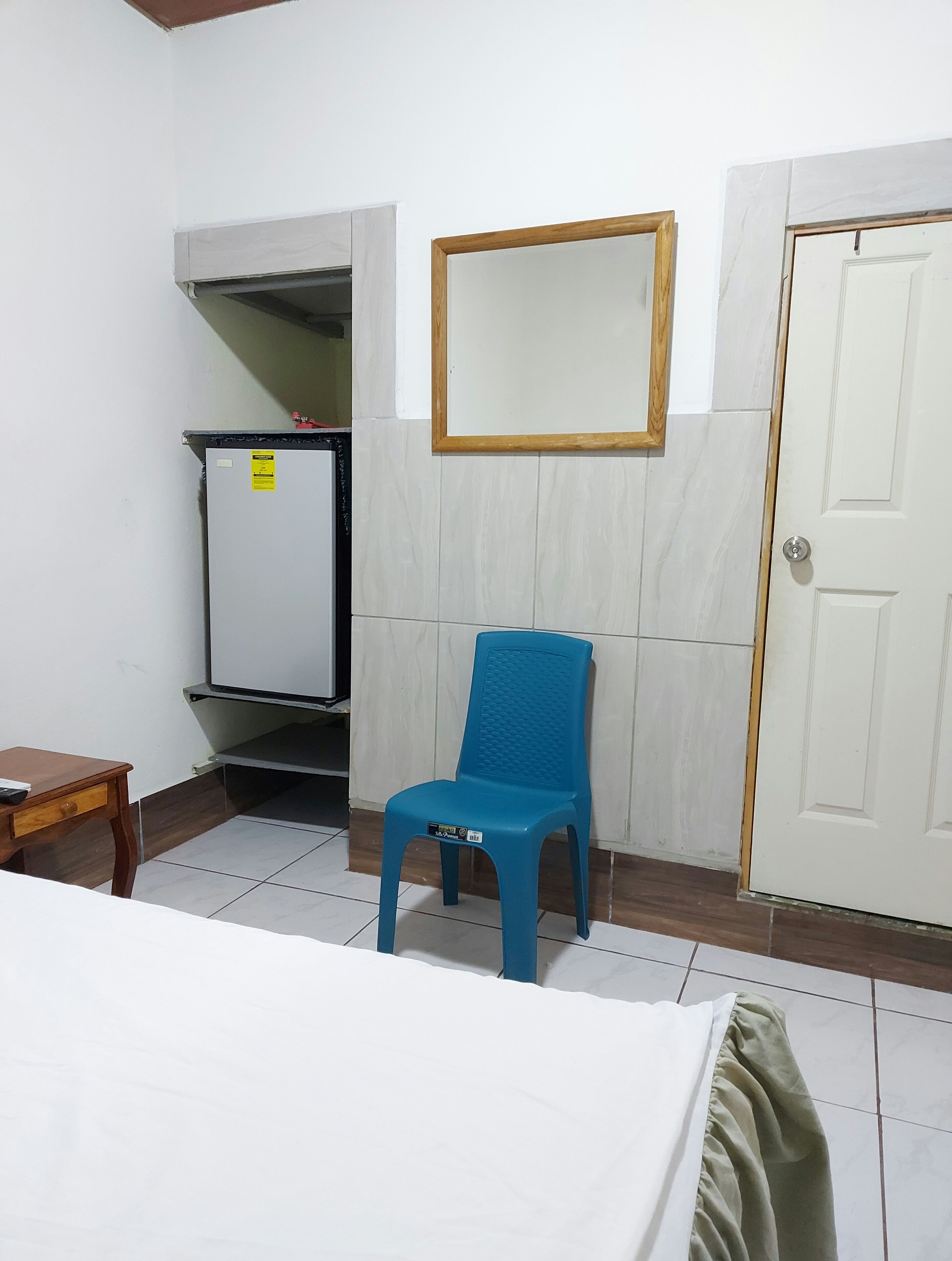 A small, clean room with a bed, a blue plastic chair, a white door, and a white wardrobe.