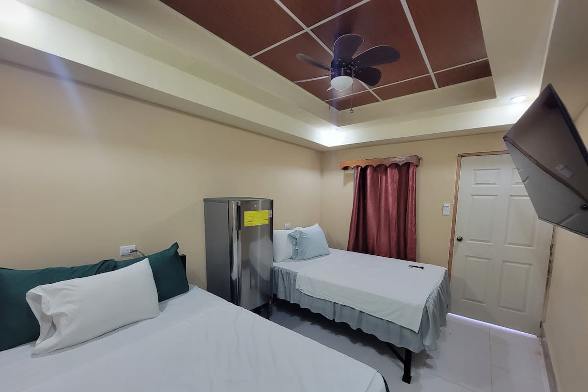Quarto de hotel com duas camas, geladeira, ventilador de teto, televisão e porta.