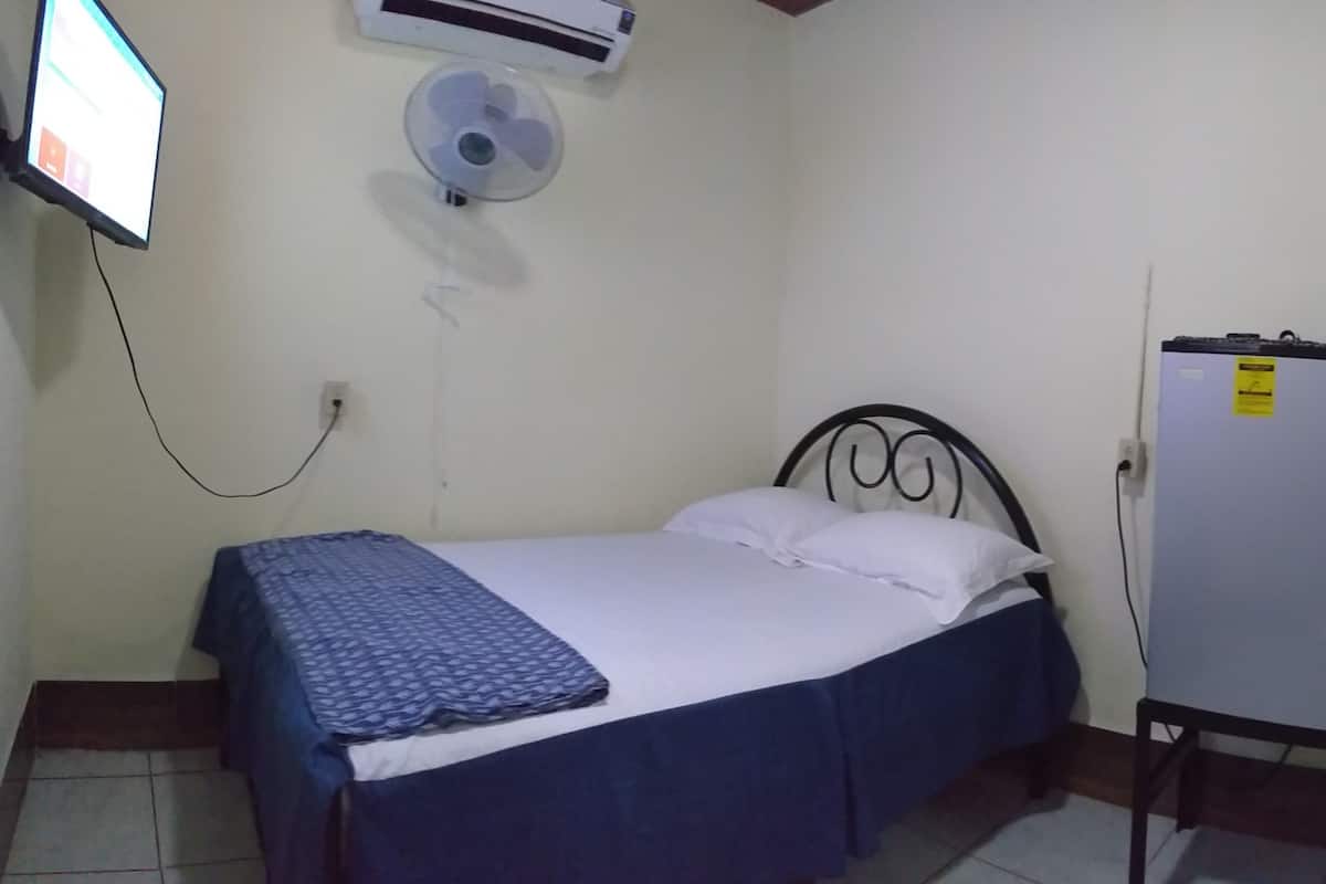 Quarto pequeno e simples com cama, ar-condicionado na parede, ventilador de teto e geladeira.