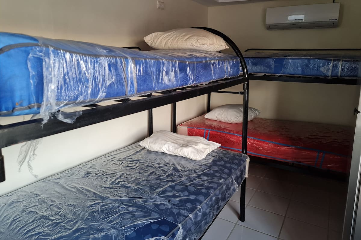 Quarto com beliche, colchão azul no colchão de cima e colchão vermelho no de baixo. Há um travesseiro branco no colchão superior e outro branco no inferior.