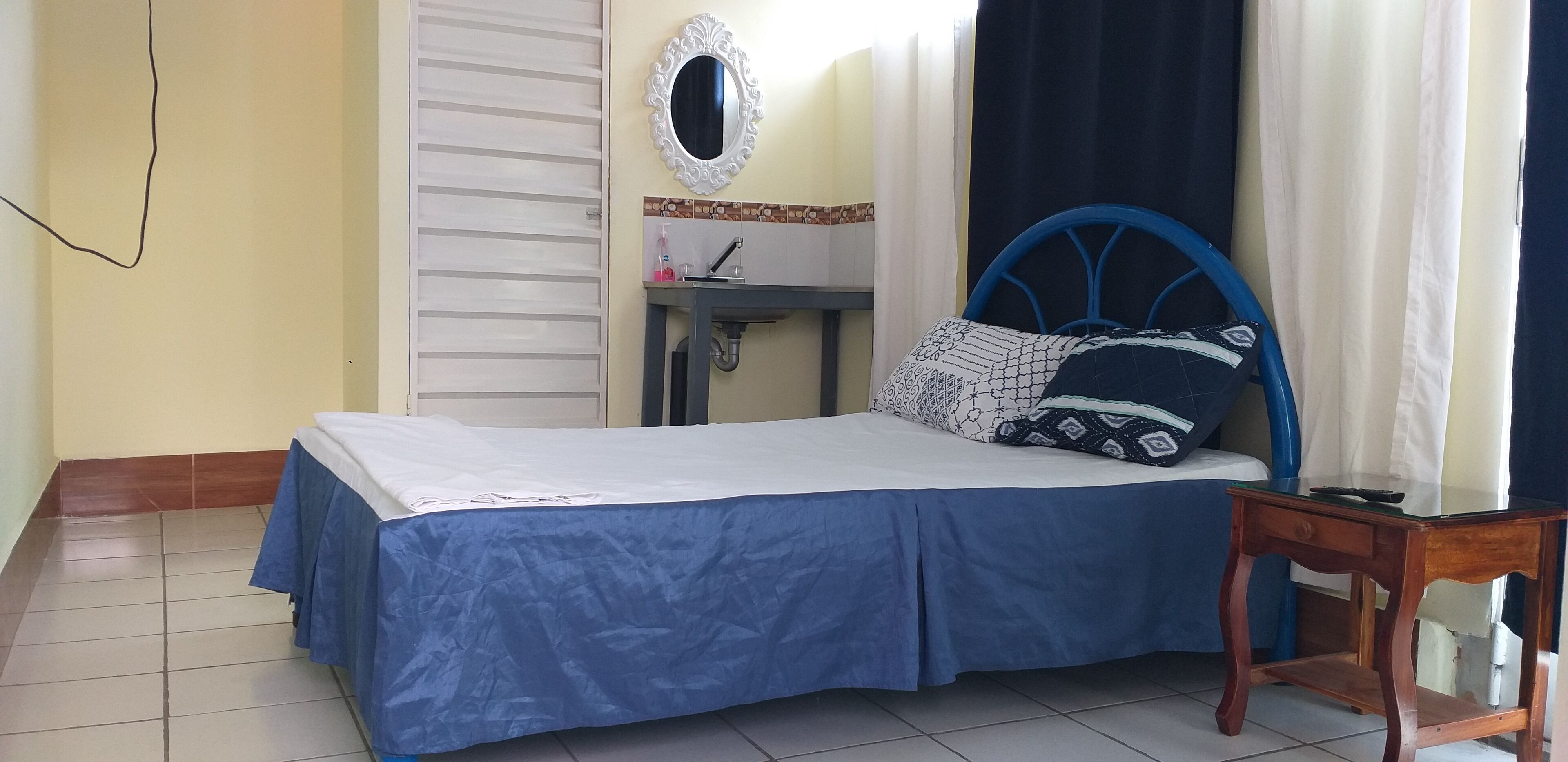 Un dormitorio con cama, mesita de noche, espejo, lavamanos y ventana con cortinas.