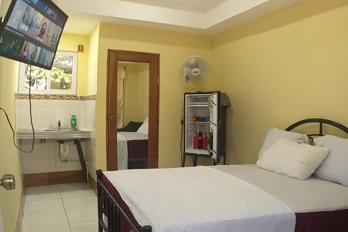 Quarto com cama, televisão na parede, pequeno espaço de cozinha e banheiro visível através de uma porta aberta.
