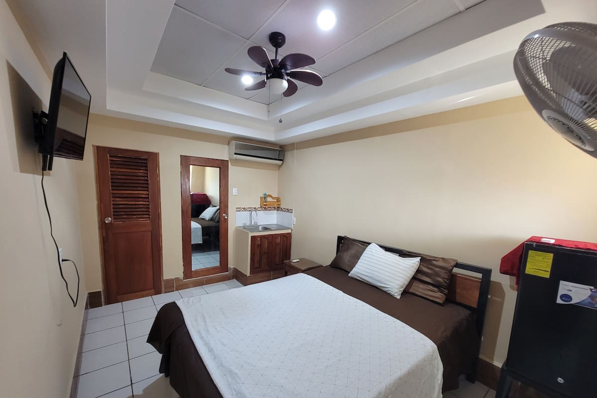 Quarto com cama, escrivaninha, cadeira, guarda-roupa, ventoinha no teto e televisão.