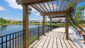 Una pérgola de madera con baranda de metal que da a un lago, con vistas a casas y un cielo despejado.