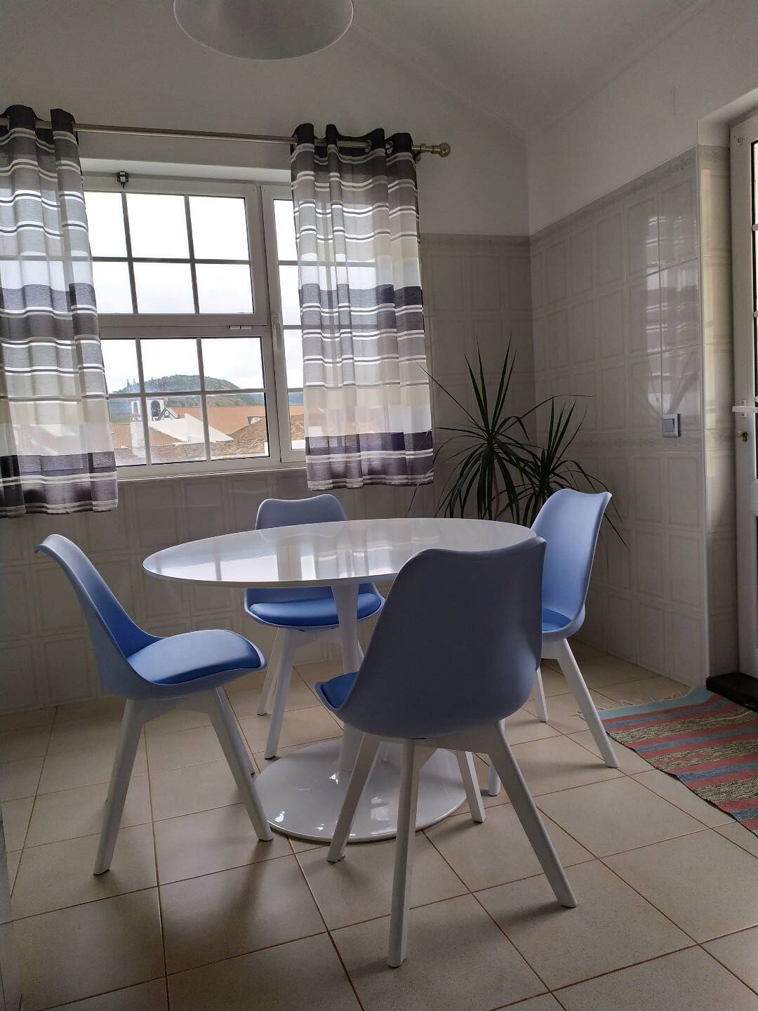 Ein Essbereich mit einem runden Tisch, vier blauen Stühlen, einer Topfpflanze und Blick ins Freie durch ein Fenster mit Vorhängen.