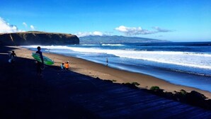 Una playa con un camino de madera, olas del océano y gente con tablas de surf.