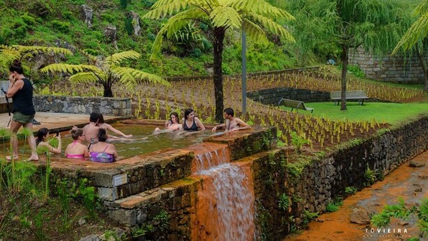 Zona de aguas termales naturales con gente relajándose en el agua, rodeada de exuberante vegetación y una cascada pequeña.