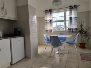 Een compacte keuken met een eethoek, inclusief een koffieapparaat, een koelkast, een ronde tafel met stoelen en een raam met gordijnen.