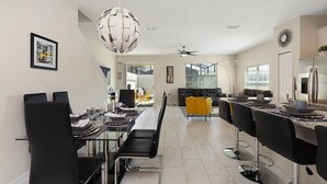 Een moderne keuken met een inbouwkoelkast, een eethoek met een glazen tafel en zwarte stoelen, en een zithoek met een bank en een geel pouf.