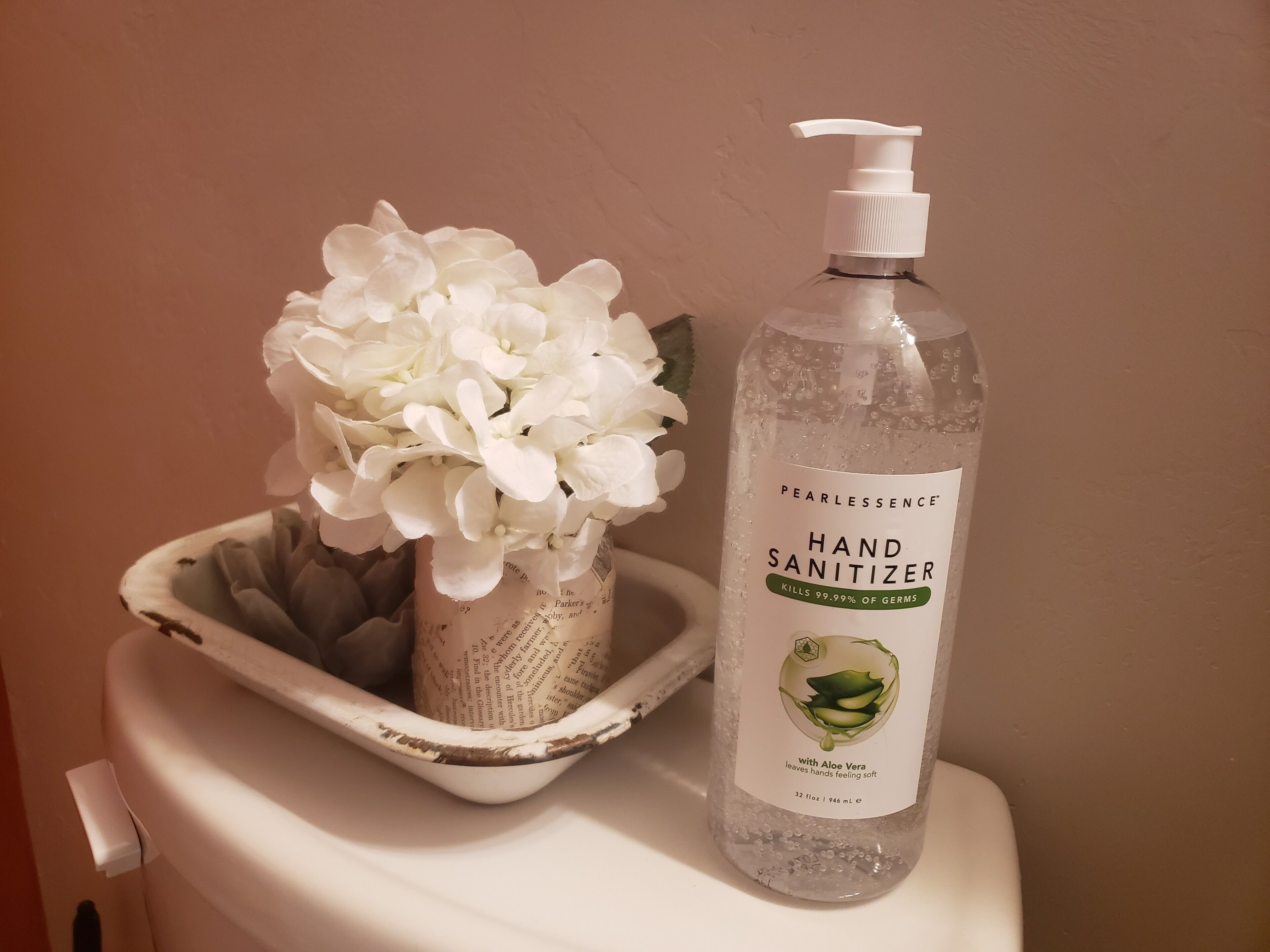 A bottle of Pearlessence hand sanitizer and a vase with white flowers on a toilet.