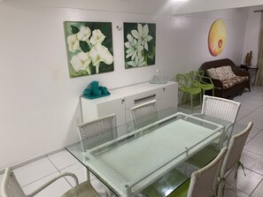A dining area with a glass table and wicker chairs, a white cabinet with teal items, and two floral paintings on the wall. - Apto. Aquaville Resort (Aquiraz)