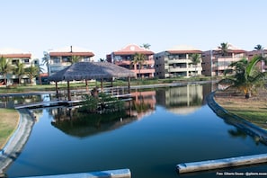A resort with a water feature, including a small pond and a bridge, surrounded by buildings with balconies and palm trees. - Apto. Aquaville Resort (Aquiraz)