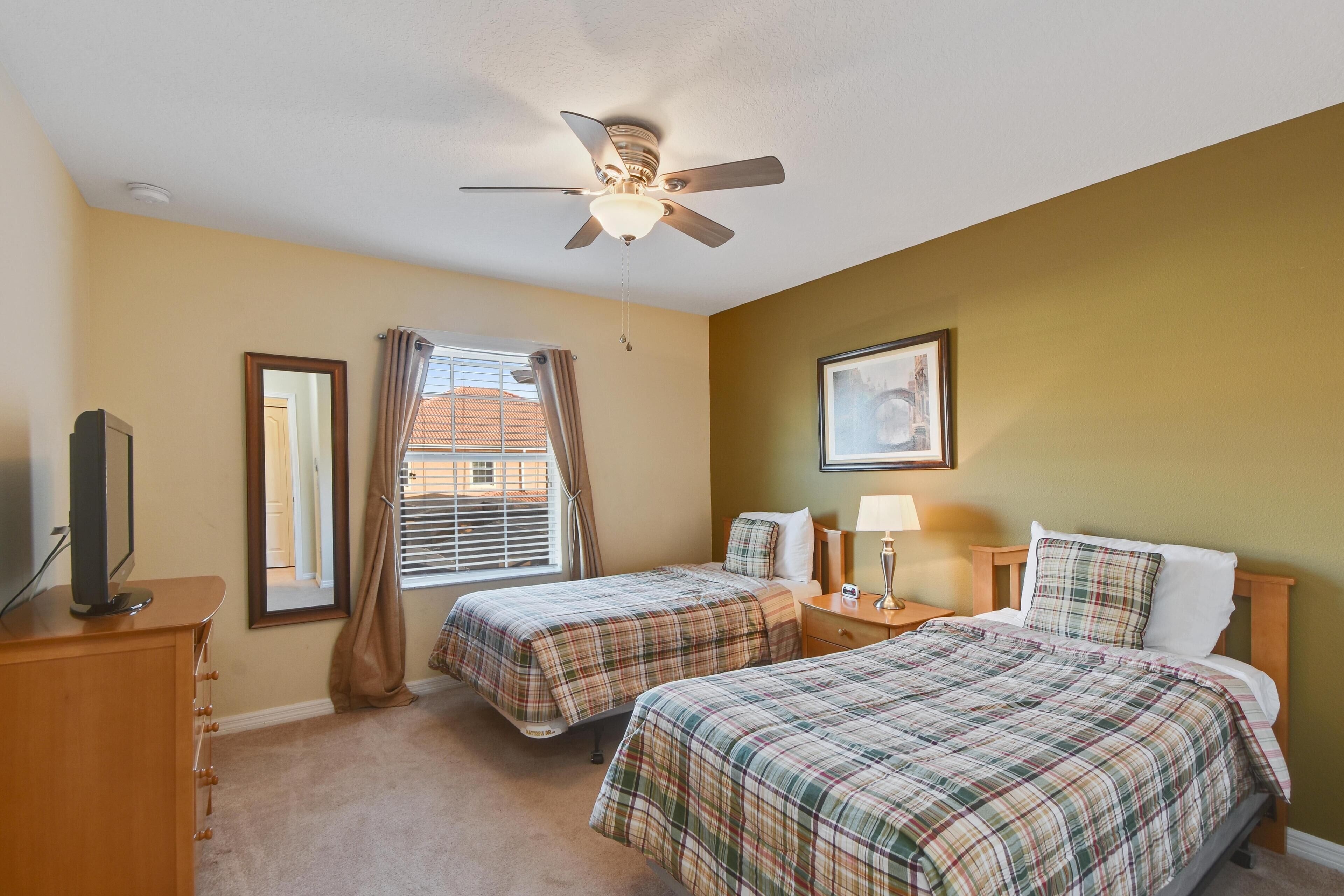 A hotel room with two beds, a ceiling fan, a television on a dresser, a window with curtains, and a framed picture on the wall.