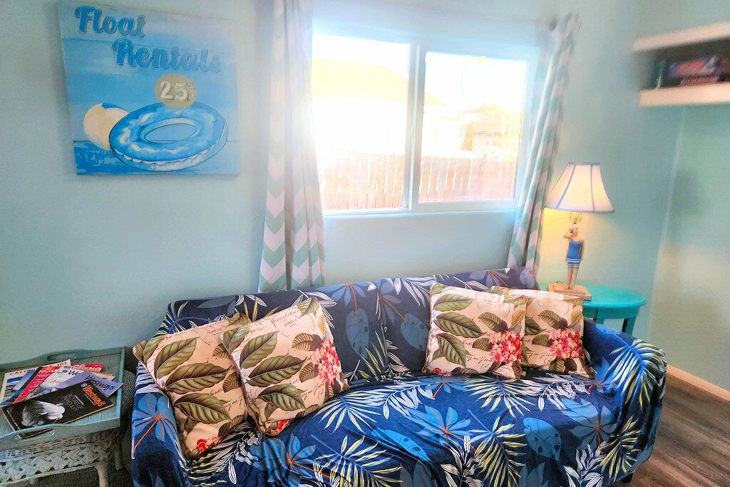 A room with a blue sofa, floral cushions, a window with curtains, a lamp, and a wall with a 'Float Rentals' sign.