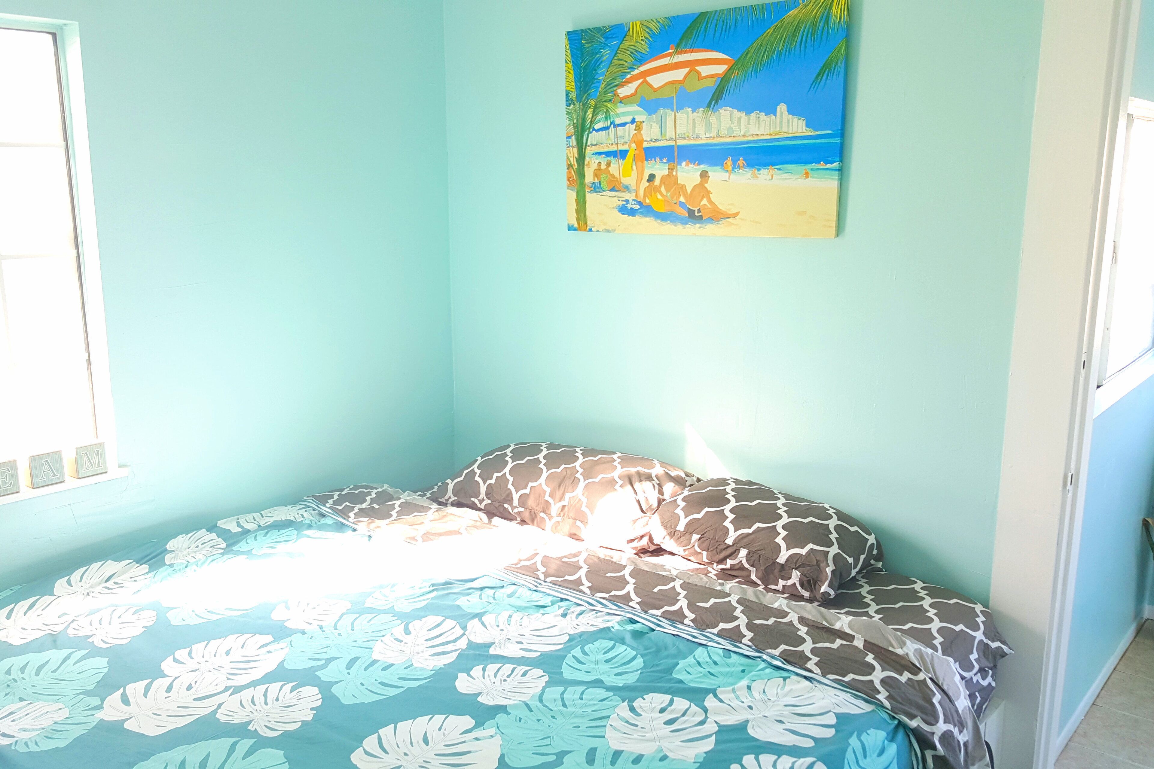 A bedroom with a bed, a painting of a beach scene, and a window.