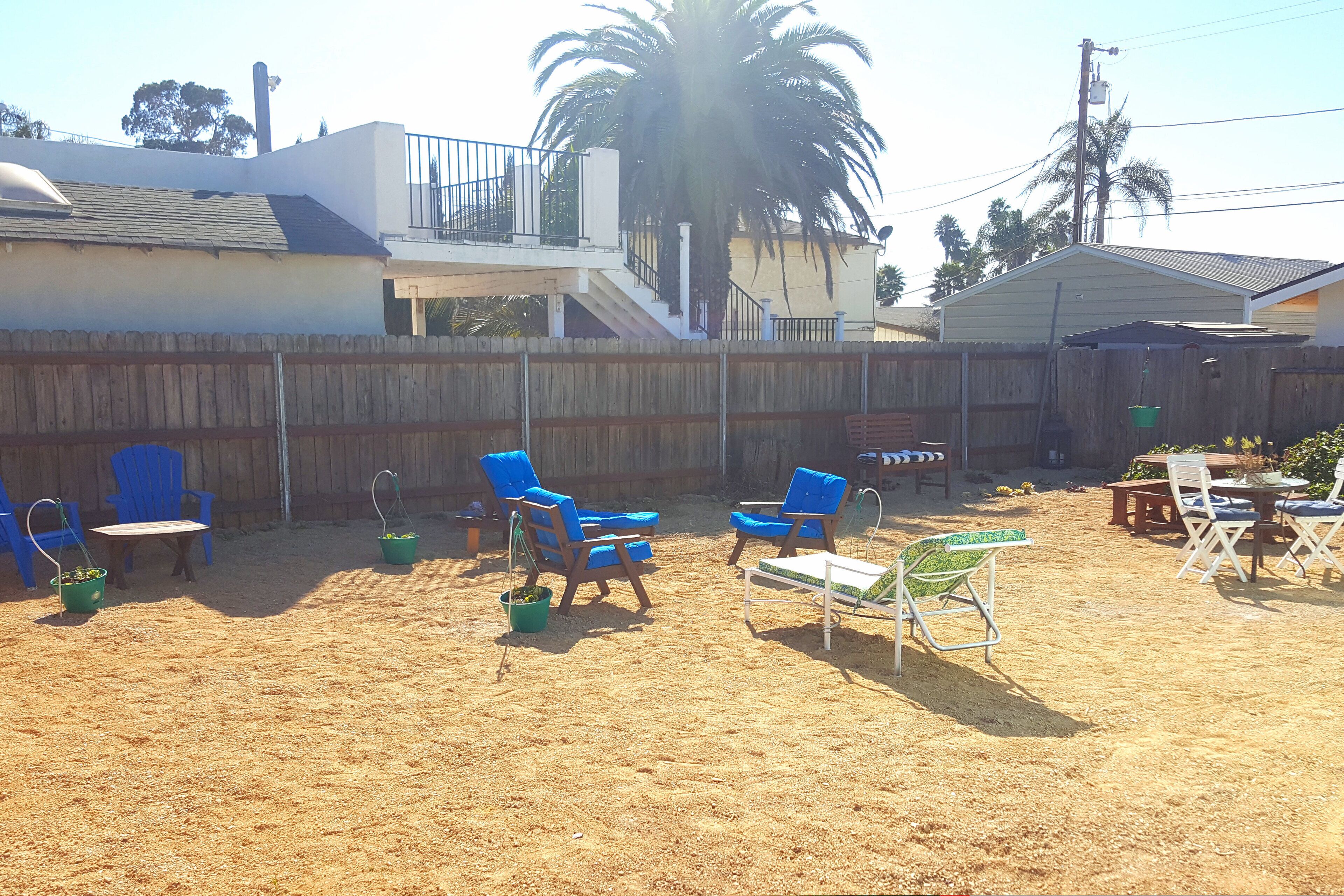 A backyard with a sandy ground, blue chairs, a table, and a palm tree.