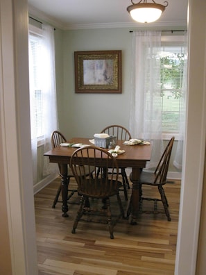 Un coin repas avec une table et des chaises en bois, un tableau encadré au mur et une fenêtre ornée de rideaux légers.