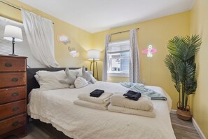 Una cama bien hecha con toallas dobladas, un aparador de madera y una planta en maceta en una habitación con paredes amarillas y una ventana con cortinas.