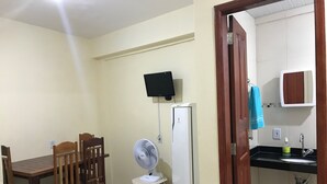 Una cocina compacta con refrigerador, un pequeño ventilador, televisor montado en la pared y una mesa de madera con sillas.