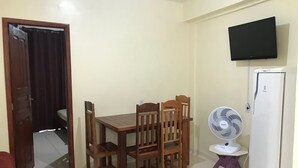 Una habitación pequeña con una mesa y sillas de madera, un refrigerador blanco, un televisor montado en la pared y un ventilador de pie.