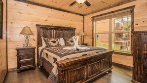 A wooden cabin bedroom with a large bed, two nightstands, a ceiling fan, and a window with a view of the outside. - Free Tickets | Theater, Game Room, Hot Tub, & Fire Pit, 1.5 Miles to Downtown (Gatlinburg)