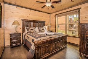 A wooden cabin bedroom with a large bed, two nightstands, a ceiling fan, and a window with a view of the outside.