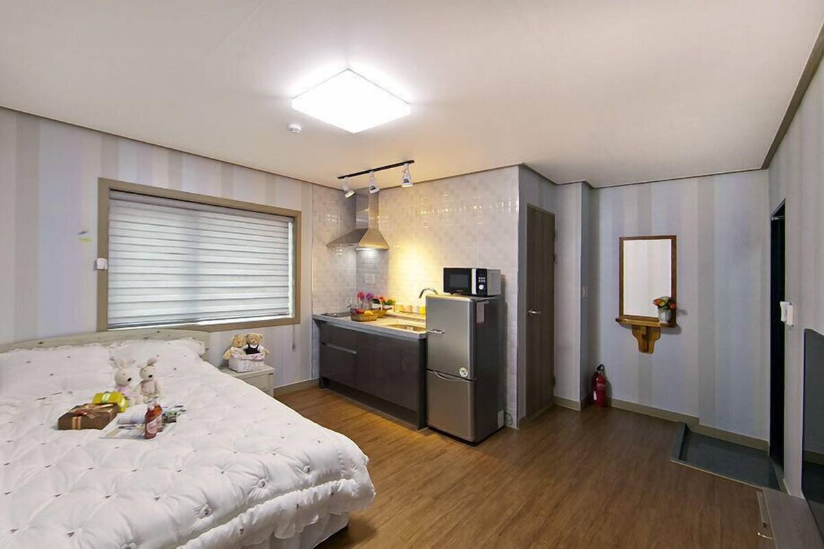 A bedroom with a bed, a small kitchenette, a bathroom with a mirror, and a window with blinds.