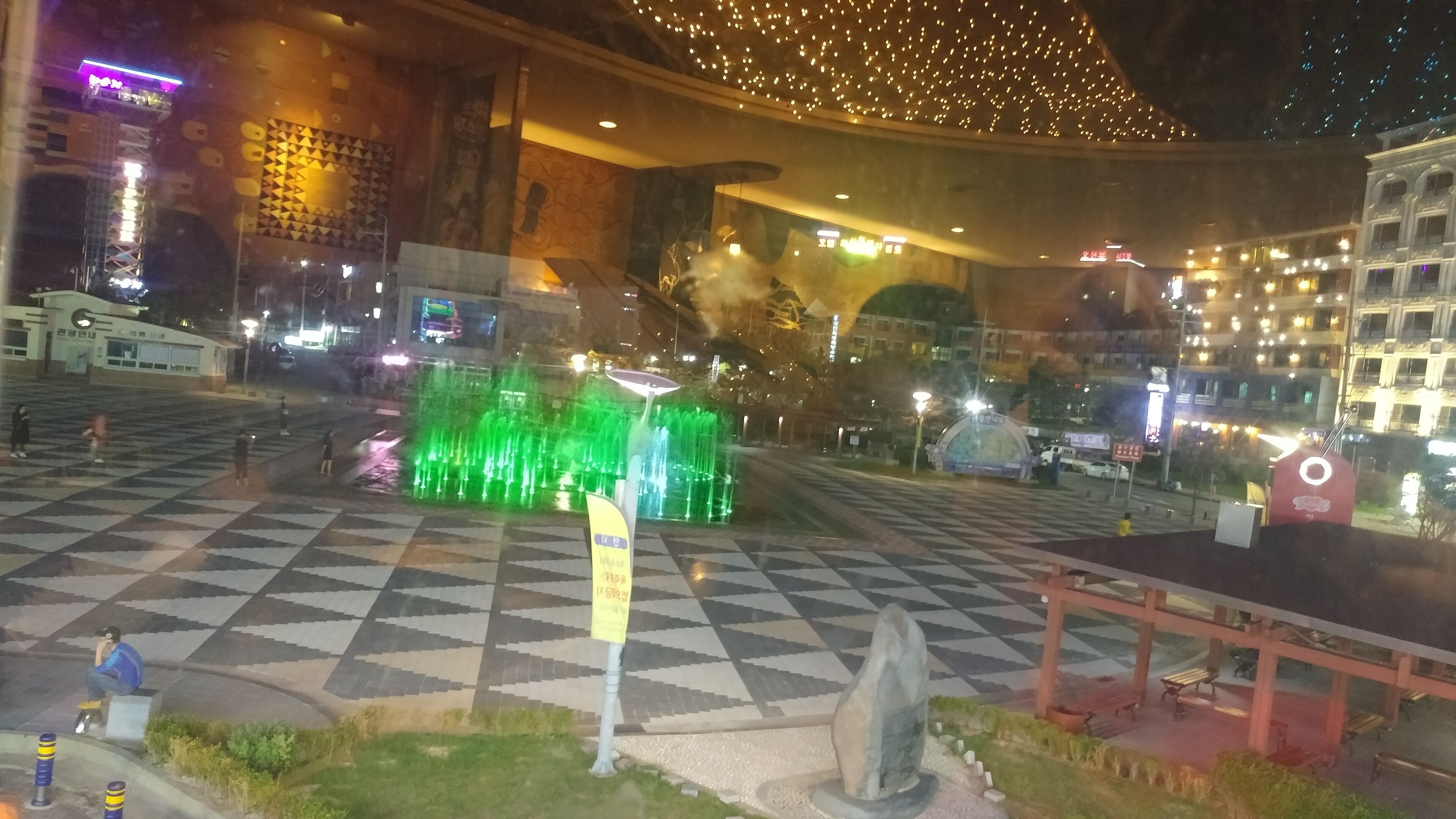 A public square with a fountain, a checkered pavement, and surrounding buildings.
