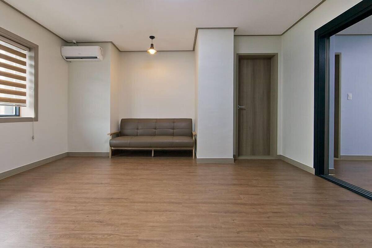 A spacious room with a wooden floor, a grey sofa, a ceiling light, and a door.