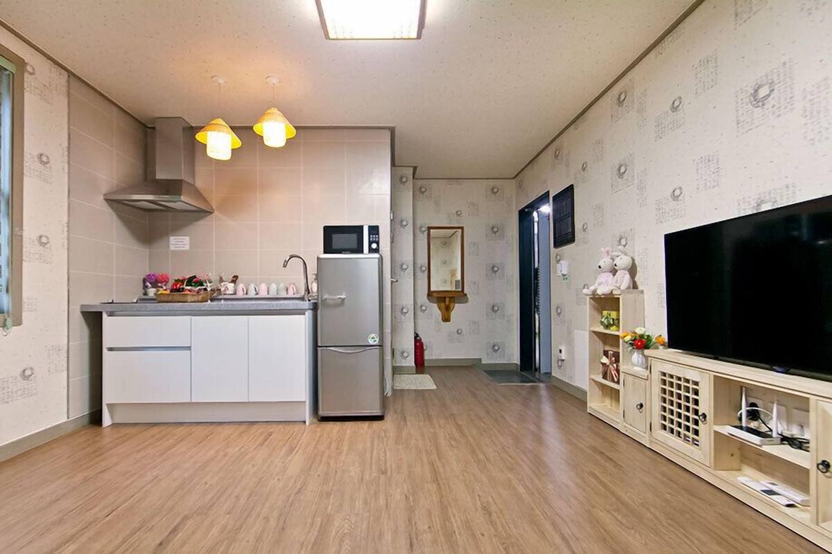 A compact kitchen with a central island, a built-in microwave, and a refrigerator. There is a small sink, a window with blinds, and a wooden cabinet with a television.