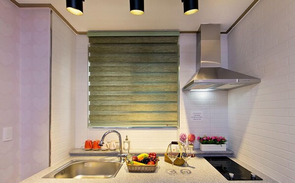 A modern kitchen with a stainless steel sink, a built-in oven, and a range hood. There is a window with blinds and a small countertop with a fruit basket and a wine glass. - Boryeong Laon Pension (Boryeong)