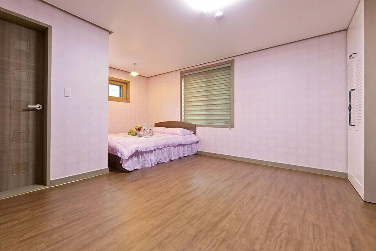 A bedroom with a wooden floor, a bed with a pink bedspread, a window with blinds, and a door with a handle.