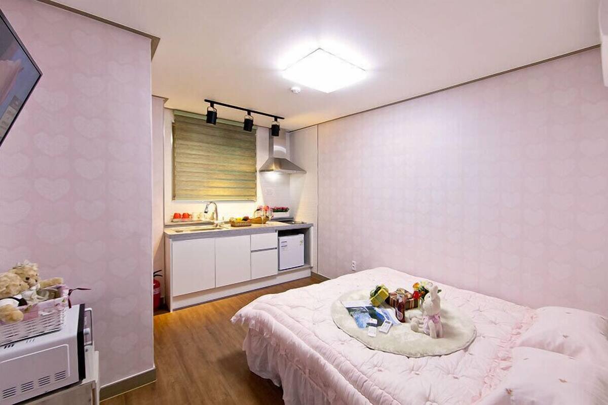 A bedroom with a bed, a bedside table, a small desk, and a kitchenette in the background.