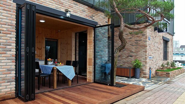 An outdoor dining area with a wooden table and chairs, a brick wall, and a glass door leading inside. - Boryeong Laon Pension (Boryeong)