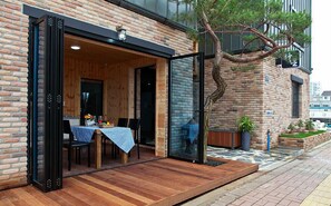 An outdoor dining area with a wooden table and chairs, a brick wall, and a glass door leading inside. - Boryeong Laon Pension (Boryeong)